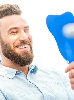 A bearded man looking at his smile in the mirror at the dentist’s office
